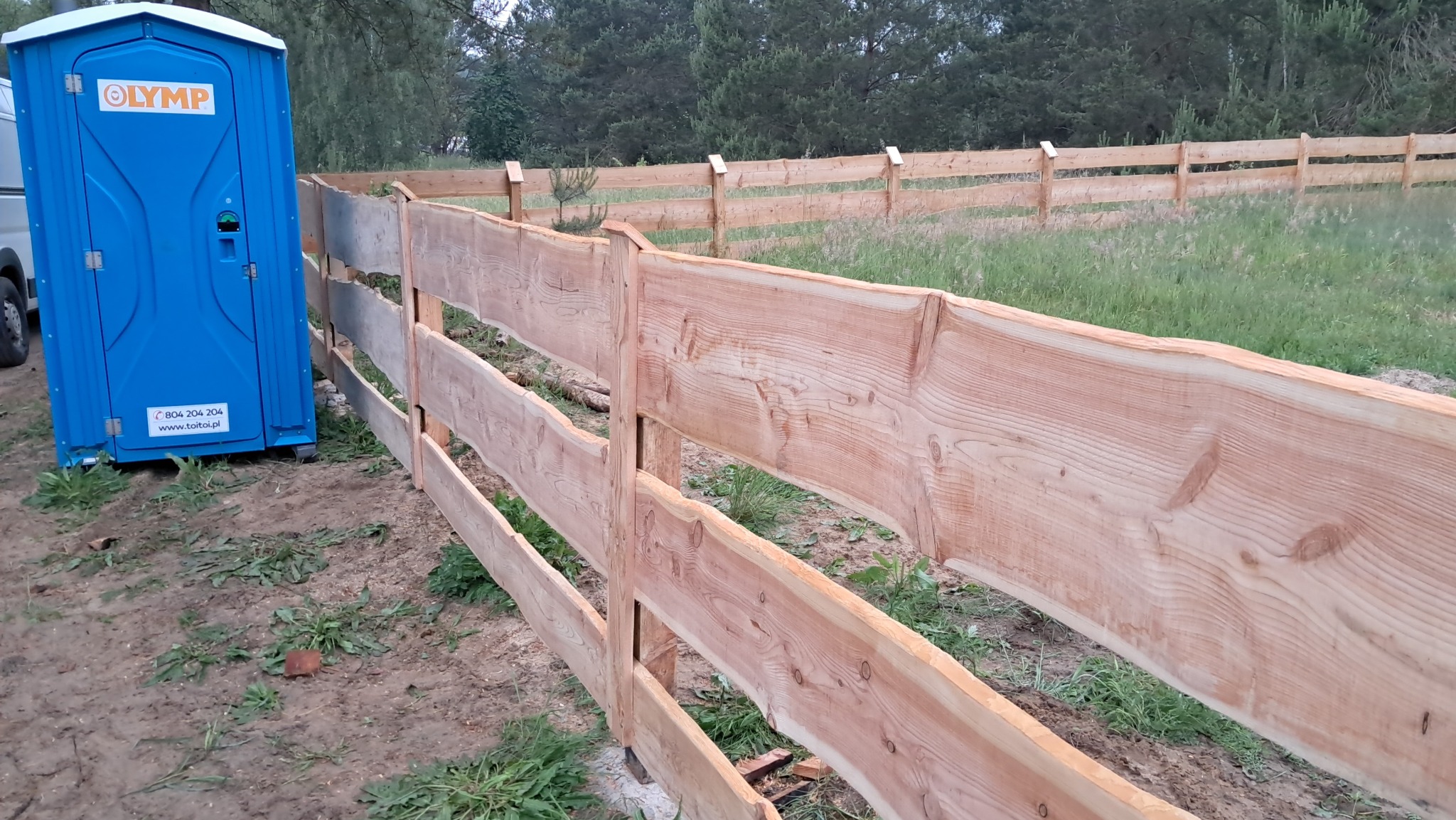 Surowe drewniane ogrodzenie z nieregularnymi krawędziami, obok przenośna toaleta w kolorze niebieskim. W tle widoczne drzewa i trawa.