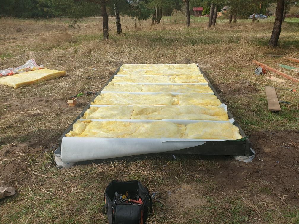 Izolacja termiczna podłogi w konstrukcji szkieletowej. Widoczne narzędzia i fragmenty konstrukcji drewnianej. Trawa i drzewa w tle. Prace w toku.