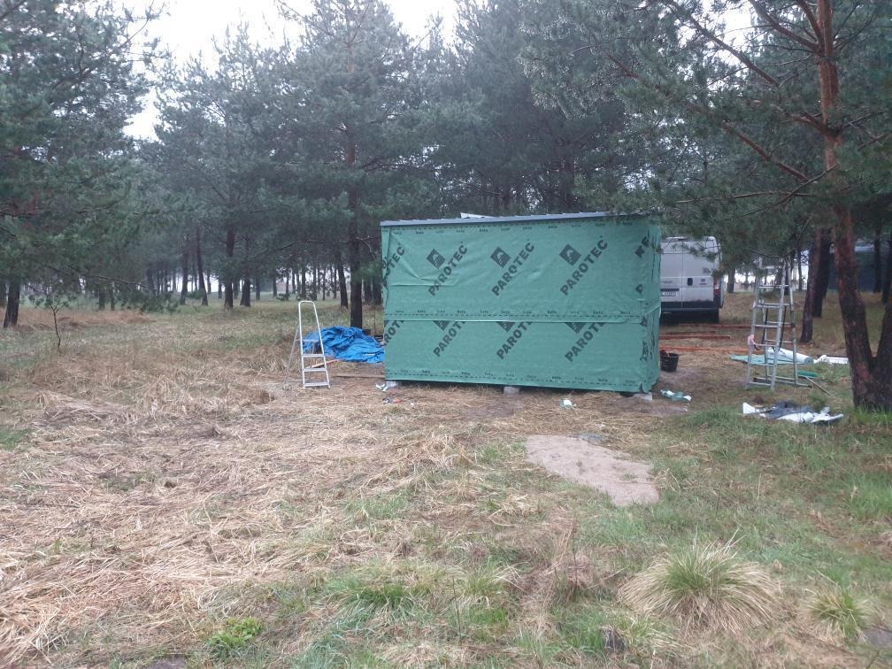 Budowa domku szkieletowego w lesie; zielona izolacja Parotec, drabina i bus w tle. Surowy krajobraz, etap wznoszenia konstrukcji.