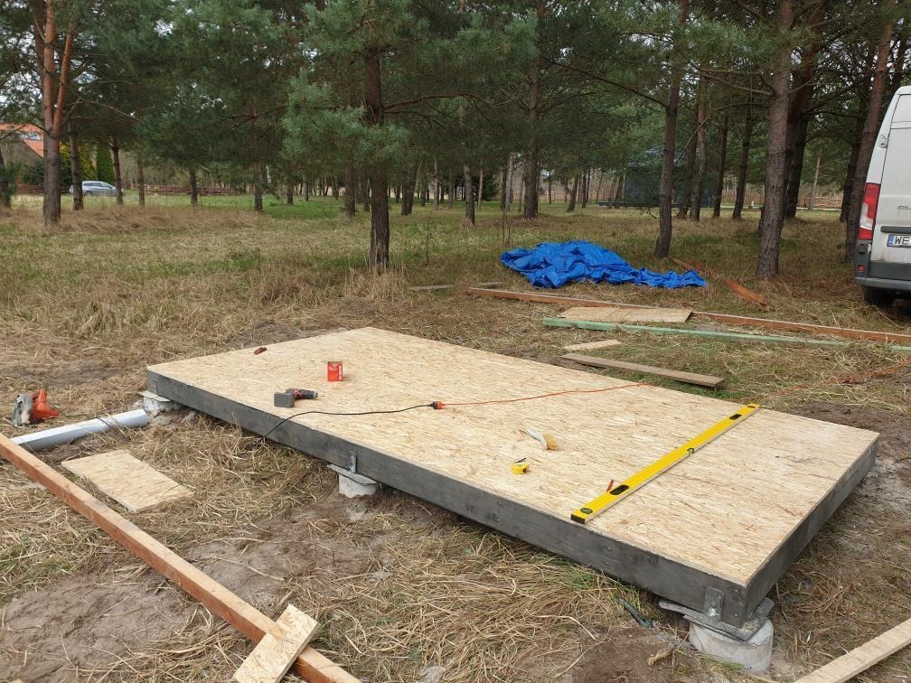 Wstępny etap budowy domu szkieletowego: przygotowany fundament z płytą OSB na betonowych wspornikach, otoczony narzędziami i elementami konstrukcyjnymi.