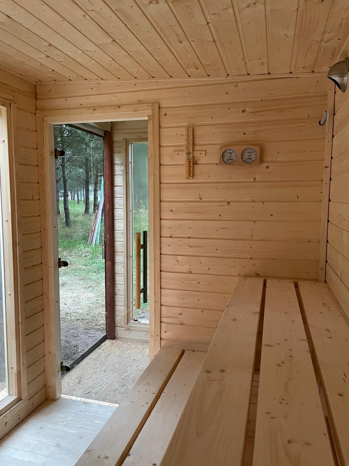 Wnętrze sauny z jasnego drewna: ławy, termometry i widok na zewnątrz przez przeszklone drzwi. Naturalne oświetlenie, surowy klimat skandynawski.