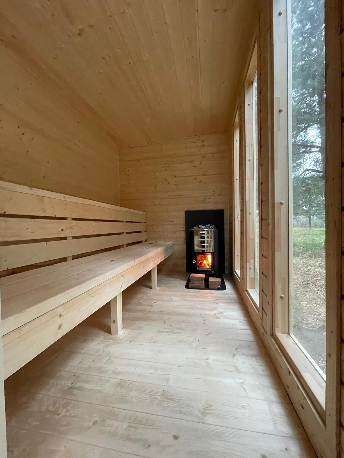 Wnętrze sauny z drewnianymi ławkami i piecem opalanym drewnem. Widok na zewnątrz przez wysokie okna. Naturalne oświetlenie.