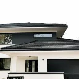 Nowoczesny dom z płaskim dachem pokrytym czarną dachówką, widoczne okna z czarnymi ramami i brama garażowa w kolorze antracytowym, biała elewacja.