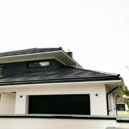 Nowoczesny dom z dwuspadowym dachem pokrytym ciemną dachówką Domino i ukrytą rynną, widoczne elementy obróbki blacharskiej oraz fragment elewacji w kolorze białym i szarym.