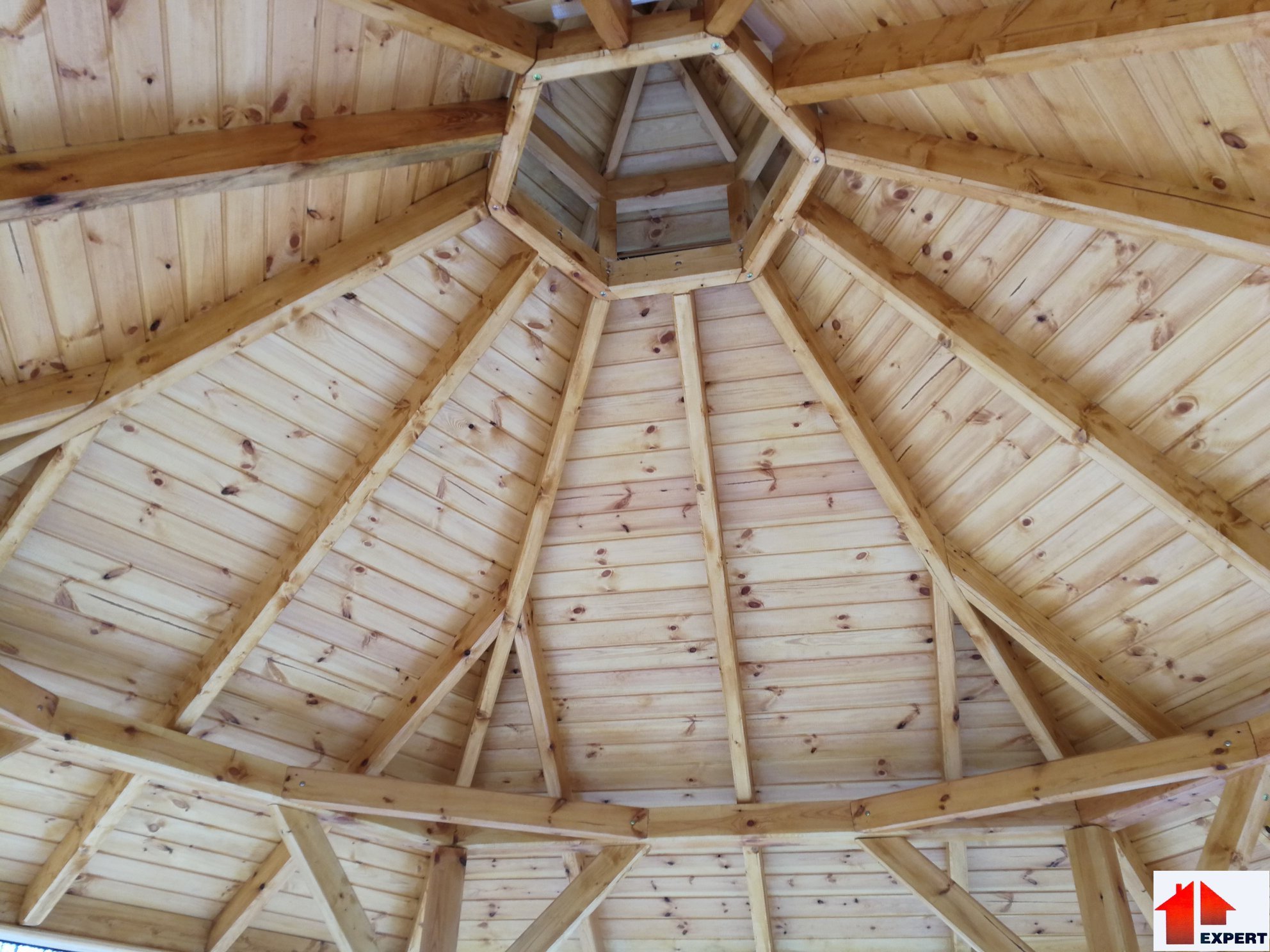 Wnętrze drewnianej altany z widoczną konstrukcją dachu pokrytą boazerią, z centralnym otworem wentylacyjnym w kształcie ośmiokąta, jasne drewno, widok z dołu.