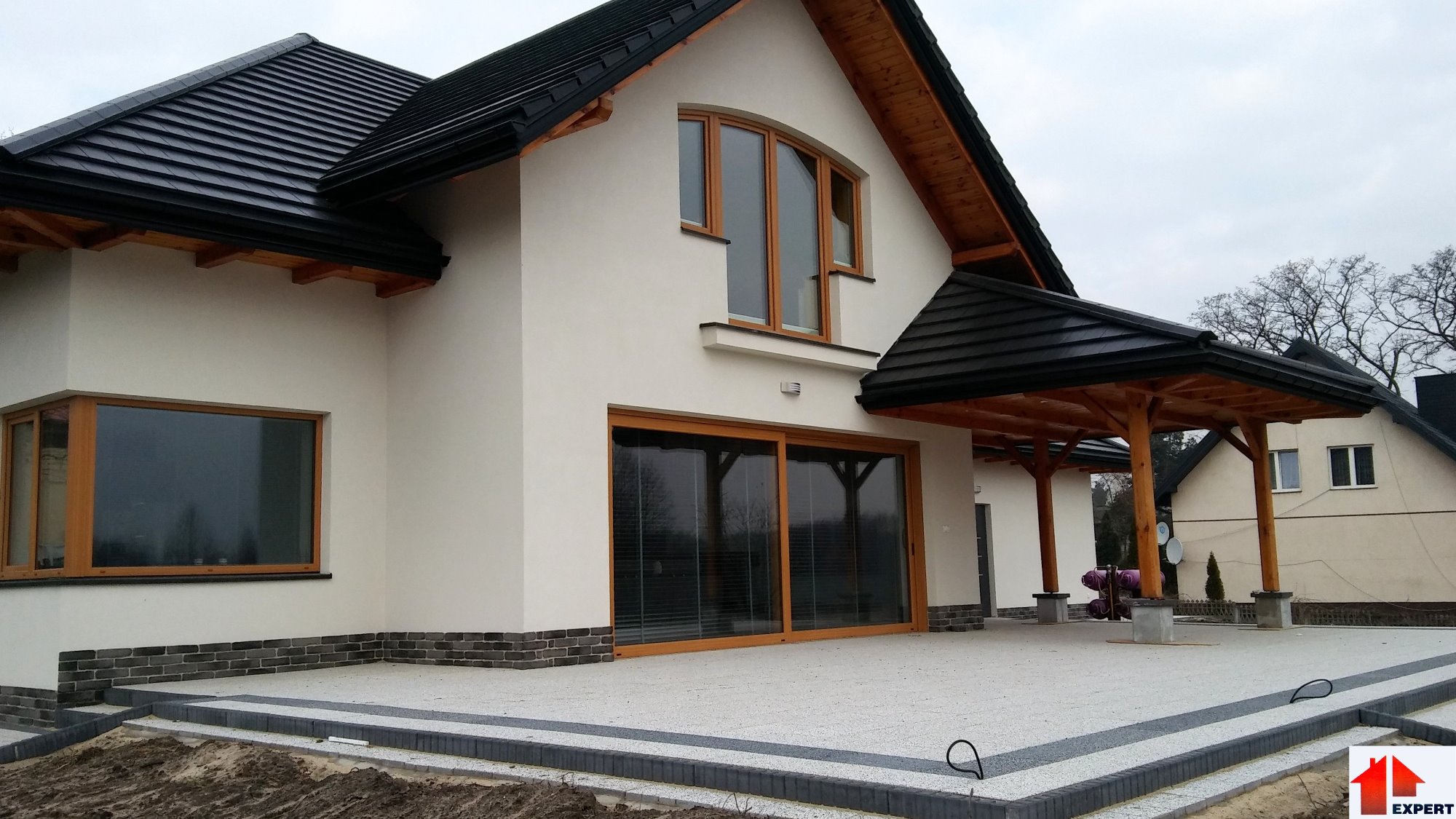 Nowoczesny dom murowany z czarną dachówką ceramiczną, drewnianą podbitką z ozdobnymi belkami i tarasem wyłożonym jasnoszarą kostką brukową.