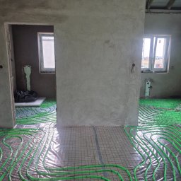 Jakub Wędzina - Zielone rury ogrzewania podłogowego ułożone na folii z siatką w surowym wnętrzu. Widoczne okna i fragmenty ścian w stanie deweloperskim.