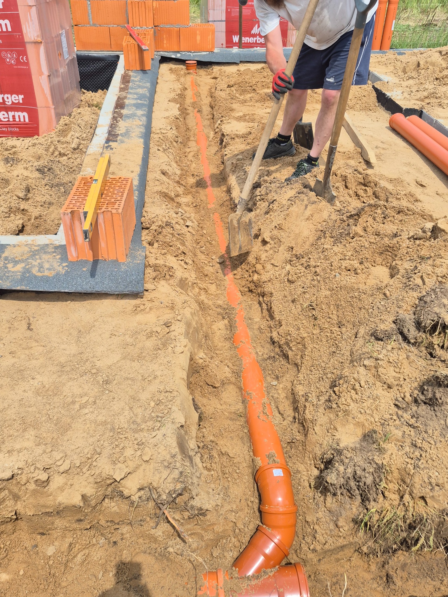 Pomarańczowe rury kanalizacyjne ułożone w wykopie, widoczne cegły i narzędzia, prace ziemne w toku. Wykop zabezpieczony, poziomica na cegłach.