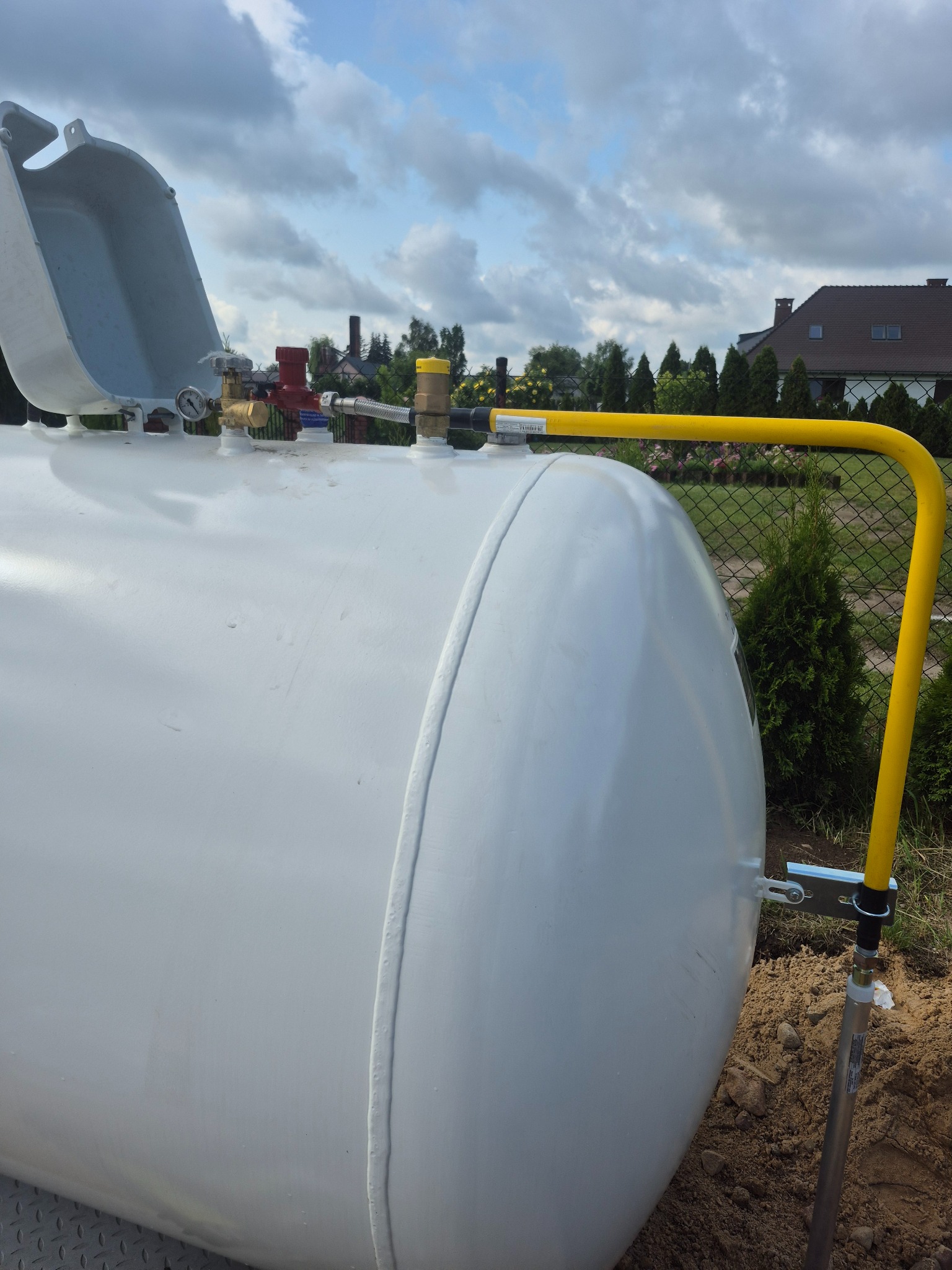 Biały zbiornik gazu płynnego z żółtą rurą doprowadzającą, widoczne zawory i manometr, na tle zieleni i błękitnego nieba z chmurami.