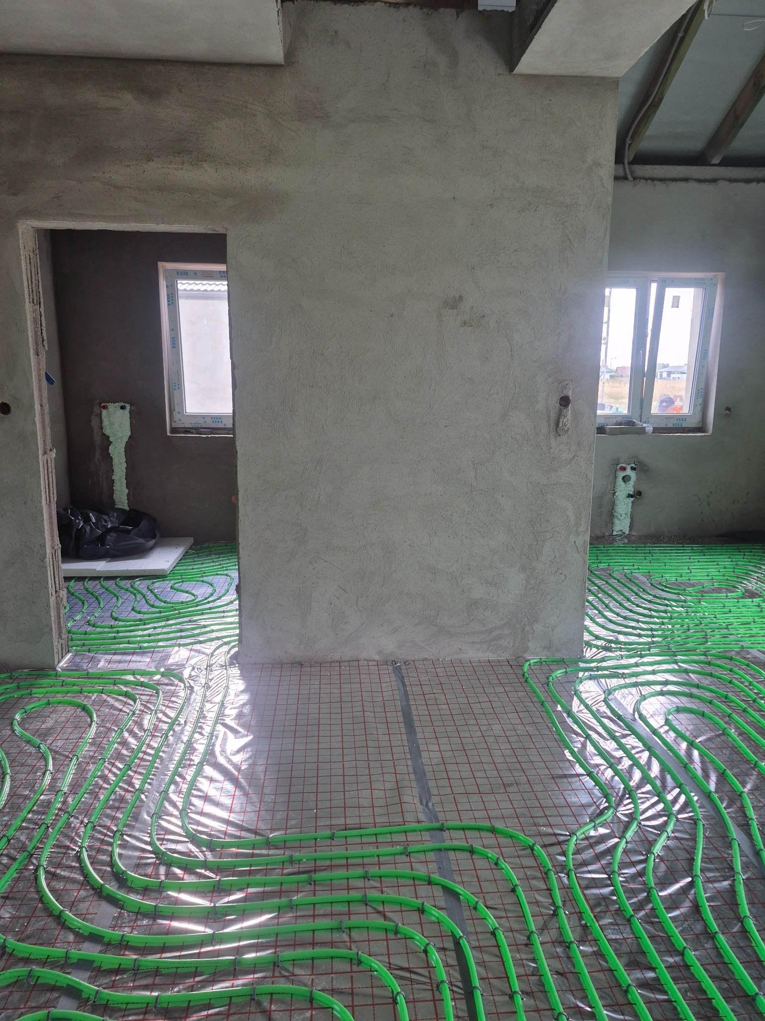 Zielone rury ogrzewania podłogowego ułożone na folii z siatką w surowym wnętrzu. Widoczne okna i fragmenty ścian w stanie deweloperskim.