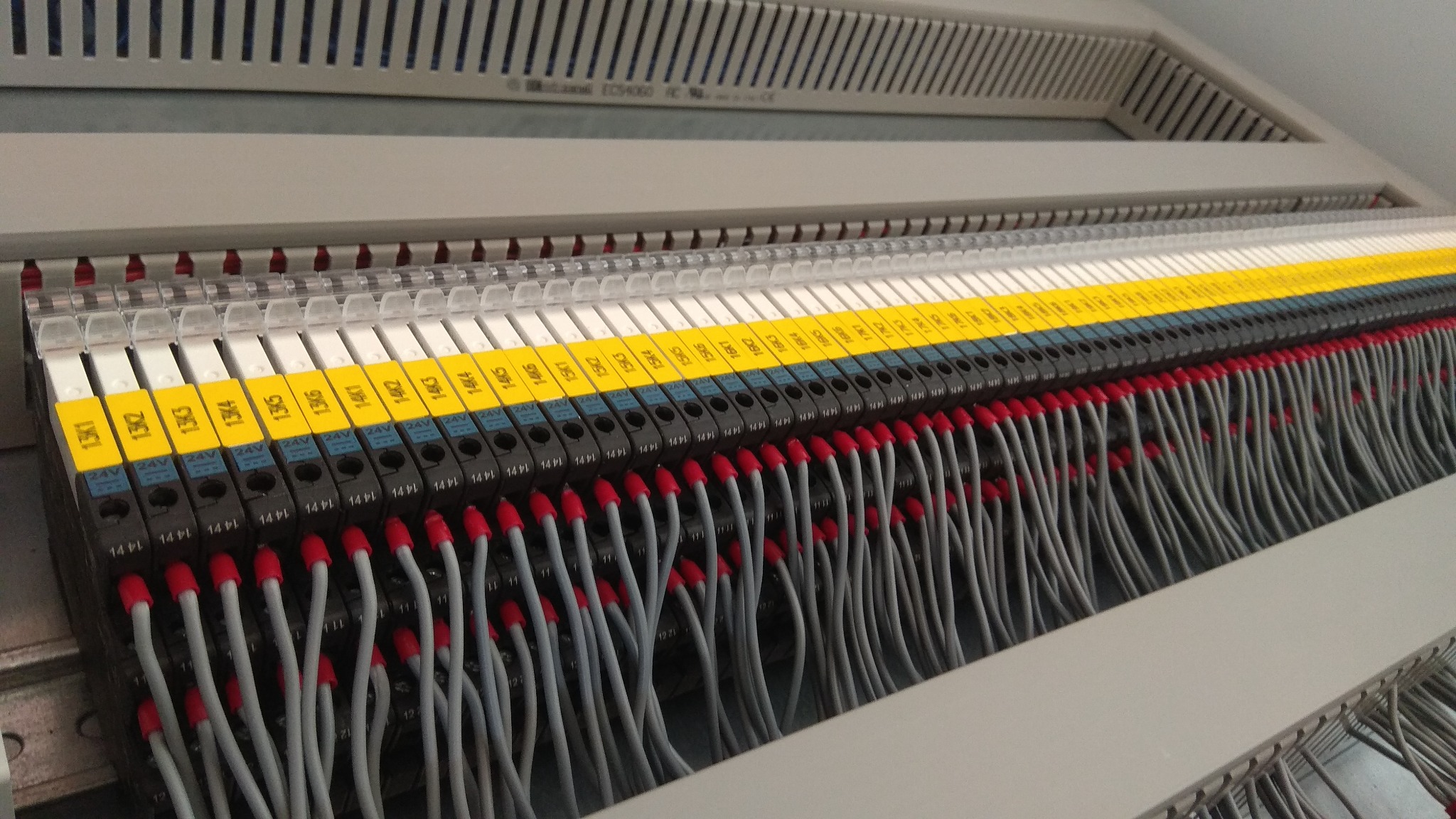 Szczegółowe ujęcie panelu sterowniczego z rzędami czarnych złączek oznaczonych żółtymi etykietami i podłączonych szarymi przewodami z czerwonymi końcówkami, widoczne oznaczenia 24V i numery...