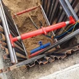 Wykop zabezpieczony metalowymi szalunkami, widoczny fragment niebieskiej rury z zaworem oraz kable instalacyjne w piasku.