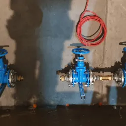 Trzy niebieskie zawory kulowe zamontowane na ścianie z betonu, połączone rurami, z widocznym czerwonym kablem zwisającym z góry.