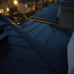 Ciemny dach z blachy na tle miejskiego krajobrazu o zmierzchu, widoczne budynki mieszkalne i oświetlenie uliczne.