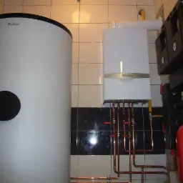 Biały zasobnik ciepłej wody marki Vaillant z czarnym panelem rewizyjnym obok wiszącego kotła gazowego tej samej marki, widoczne miedziane rury i czerwony zbiornik wyrównawczy.