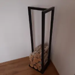 Nowoczesny, minimalistyczny stojak na drewno kominkowe wykonany z czarnego metalu, wypełniony pociętymi brzozowymi klockami, ustawiony w rogu pomieszczenia z drewnianą podłogą.