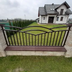 Nowoczesne ogrodzenie metalowe z dekoracyjnymi łukami, osadzone na murku z kamienia łupanego, na tle domu z ciemną dachówką i rzędu tuj.
