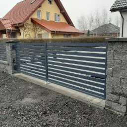 Nowoczesna, przesuwna brama wjazdowa z poziomych, szarych profili, osadzona na słupkach z szarej kostki betonowej, na tle domu z czerwoną dachówką i posesji wysypanej grysem.