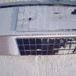 Widok z góry na instalację fotowoltaiczną częściowo zasypaną śniegiem na dachu budynku z czerwoną dachówką, otoczonego śnieżnym terenem z widocznymi śladami. Część paneli jest odsłonięta.