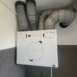 Zamontowany rekuperator ThesslaGreen AirPack4 400V Enthalpy z podłączonymi przewodami wentylacyjnymi, widoczne szare rury i biała obudowa urządzenia na tle szarej ściany.