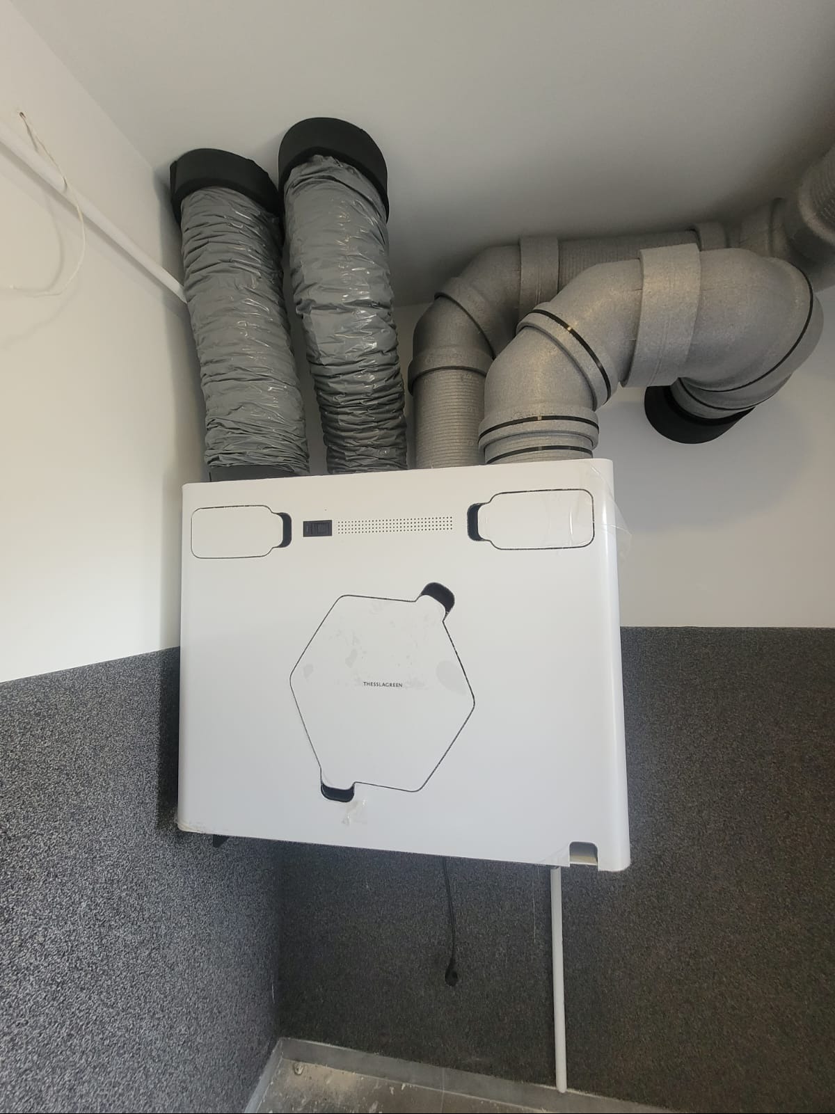 Zamontowany rekuperator ThesslaGreen AirPack4 400V Enthalpy z podłączonymi przewodami wentylacyjnymi, widoczne szare rury i biała obudowa urządzenia na tle szarej ściany.