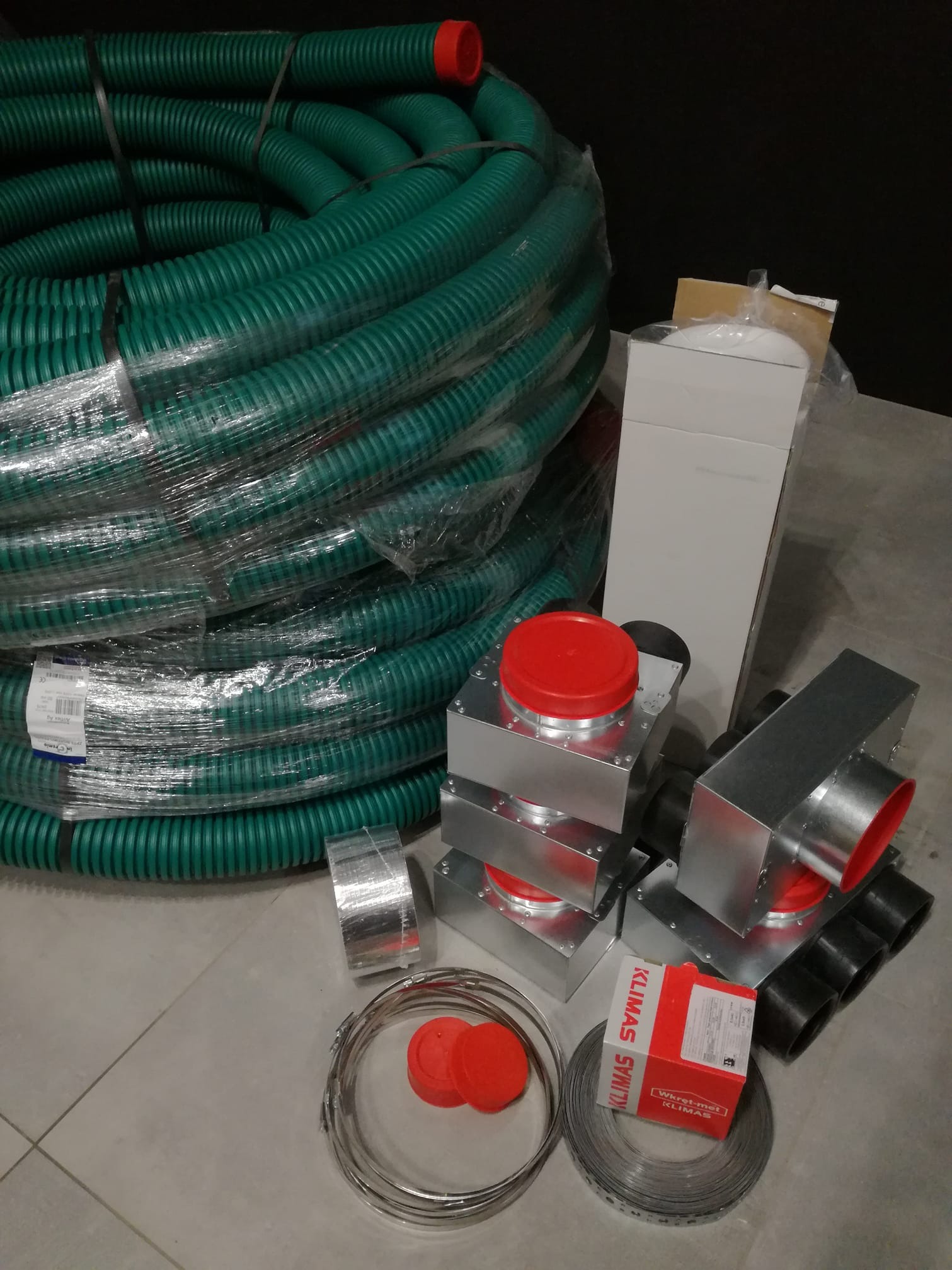 Zestaw komponentów systemu wentylacyjnego: zwój zielonego karbowanego przewodu w folii, metalowe kształtki z czerwonymi zaślepkami, taśma montażowa, pudełko Klimas-Wkretmet.