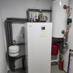 Biała pompa ciepła NIBE z zbiornikiem i orurowaniem w kotłowni. Widoczne liczniki, zawory i izolacja termiczna. Szare rury i płytki podłogowe.