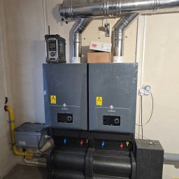 Dwa szare piece gazowe De Dietrich z podłączeniami, rurami spalinowymi i skrzynką gazową. Widoczny radiobudzik Makita. Instalacja w pomieszczeniu technicznym.