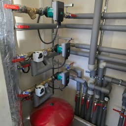 Złożona instalacja grzewcza z czerwonym zbiornikiem wyrównawczym, rurami w izolacji i pompami obiegowymi, zamontowana przy ścianie. Widoczne zawory i manometry.