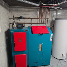 Turkusowy piec na pellet z czerwonymi drzwiczkami i zasobnik wody w kotłowni z szarymi płytkami. Widoczne rury miedziane i instalacja.
