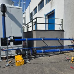 Termomax - Instalacja rur w kolorze niebieskim, przymocowanych do ściany budynku i agregatu chłodniczego. Żółta skrzynka narzędziowa i sprzęt pomiarowy na ziemi.