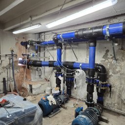 Termomax - Złożona instalacja hydrauliczna z niebieskich rur i czarnych łączników, wsparta na metalowych konstrukcjach, z dwoma niebieskimi pompami w surowym pomieszczeniu.