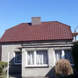 Dom jednorodzinny z nowym, brązowym dachem z blachodachówki i murowanym kominem z cegły, na tle błękitnego nieba, widoczne rusztowanie obok budynku.