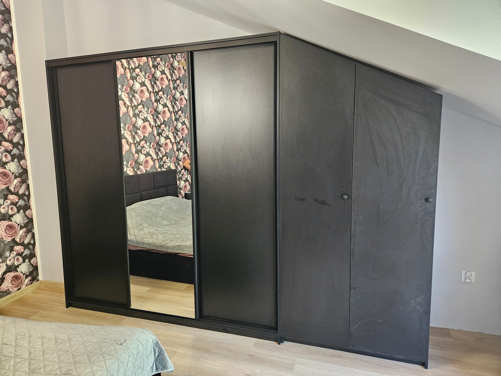 Czarna szafa wnękowa z przesuwnymi drzwiami, jedno skrzydło z lustrem odbijającym wzorzystą tapetę i fragment łóżka, dopasowana do skosu poddasza.