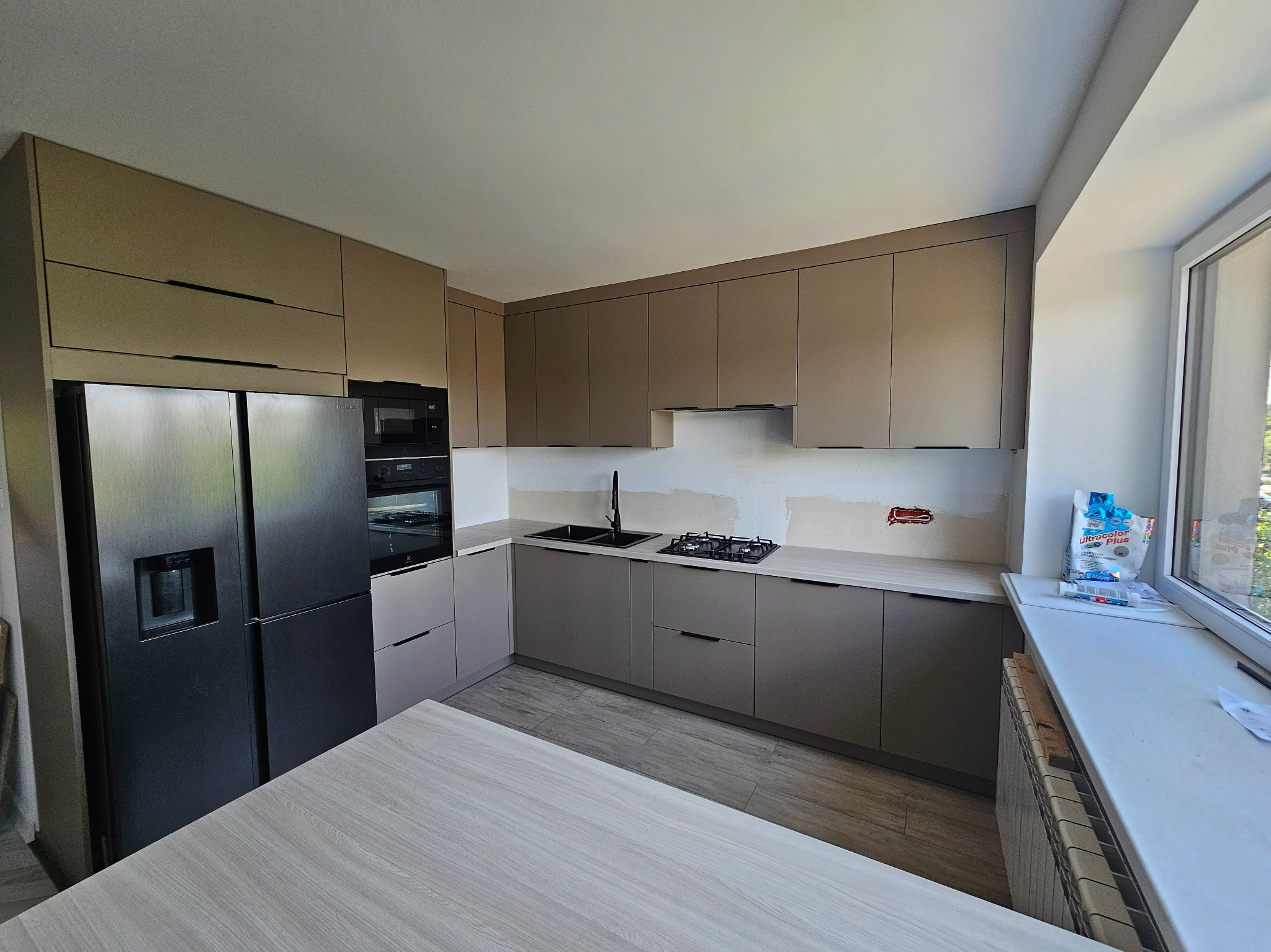 Jasna kuchnia w trakcie montażu, z widocznymi niedokończonymi pracami wykończeniowymi ścian, zabudowa w odcieniach beżu i szarości, czarny sprzęt AGD, na blacie przy oknie worek z zaprawą.