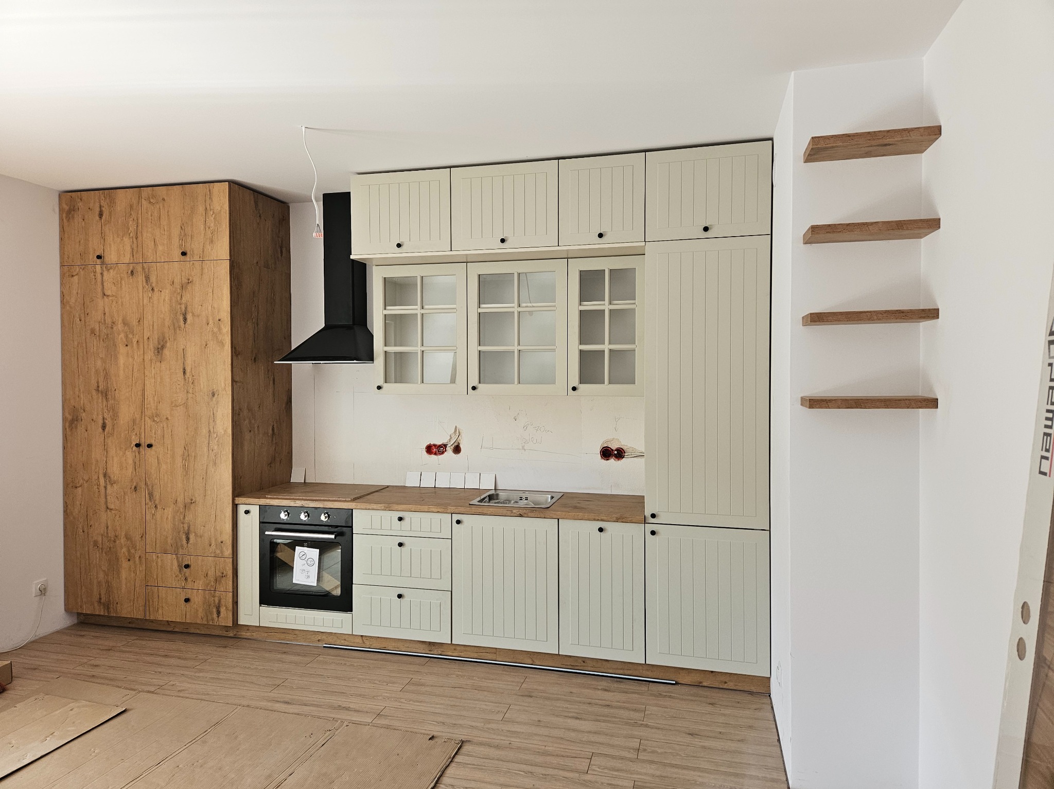 Montaż jasnej kuchni w stylu rustykalnym z drewnianymi blatami i czarnym okapem, widoczne niedokończone wykończenia ścian i podłogi, oraz drewniane półki na ścianie.