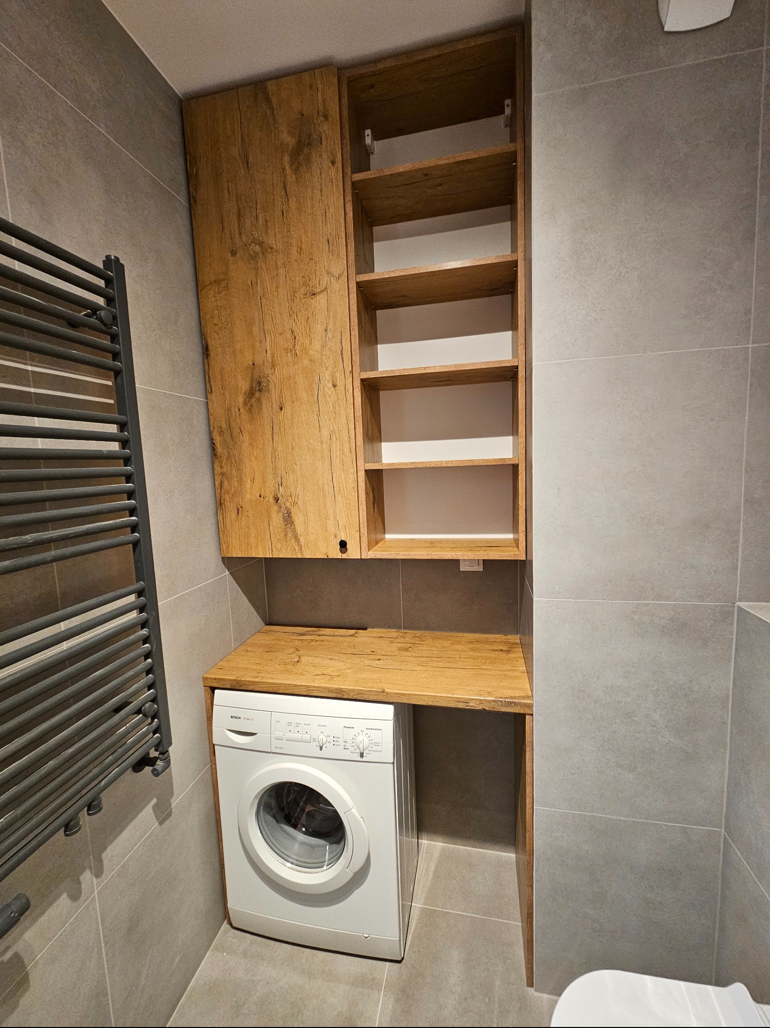 Łazienka z zabudową pralki: szafka wisząca z drewnianymi frontami i otwartymi półkami nad pralką Bosch, blat z drewna, szary grzejnik drabinkowy na ścianie z szarych płytek.
