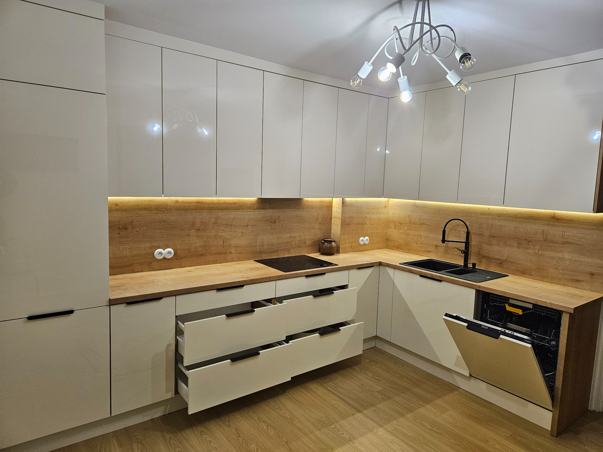 Jasna kuchnia z połyskiem, drewniany blat i oświetlenie LED pod szafkami, otwarte szuflady i zmywarka, nowoczesna lampa sufitowa.
