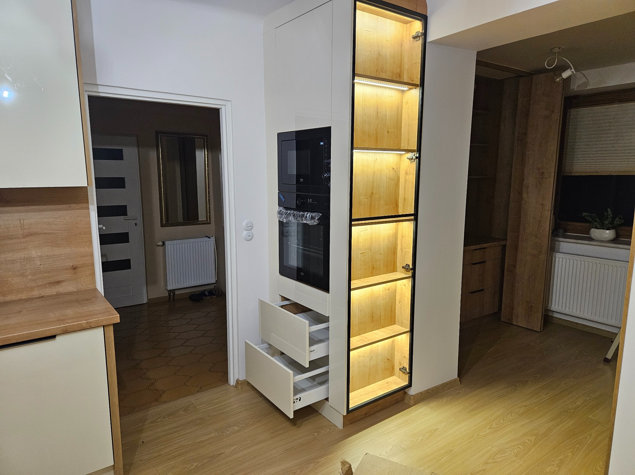 Biała zabudowa kuchenna z szufladami i piekarnikiem, oświetlona szafka z drewnianymi półkami, widoczne otwarte szuflady i drzwi do innego pomieszczenia w tle.