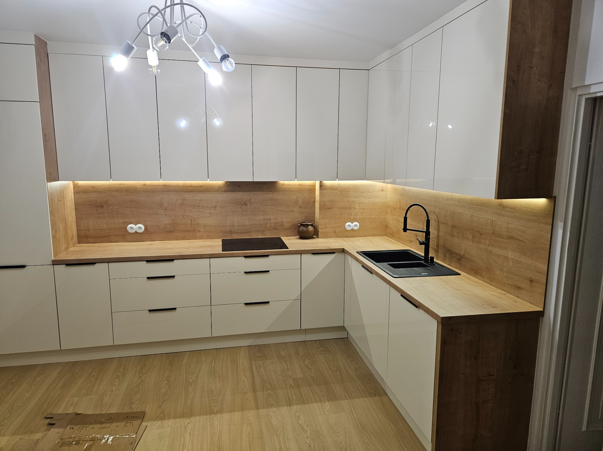 Nowoczesna kuchnia z białymi, błyszczącymi frontami szafek, drewnianym blatem i oświetleniem LED pod szafkami wiszącymi. Czarna bateria i zlew kontrastują z jasnym blatem. Na blacie widoczna płyta...