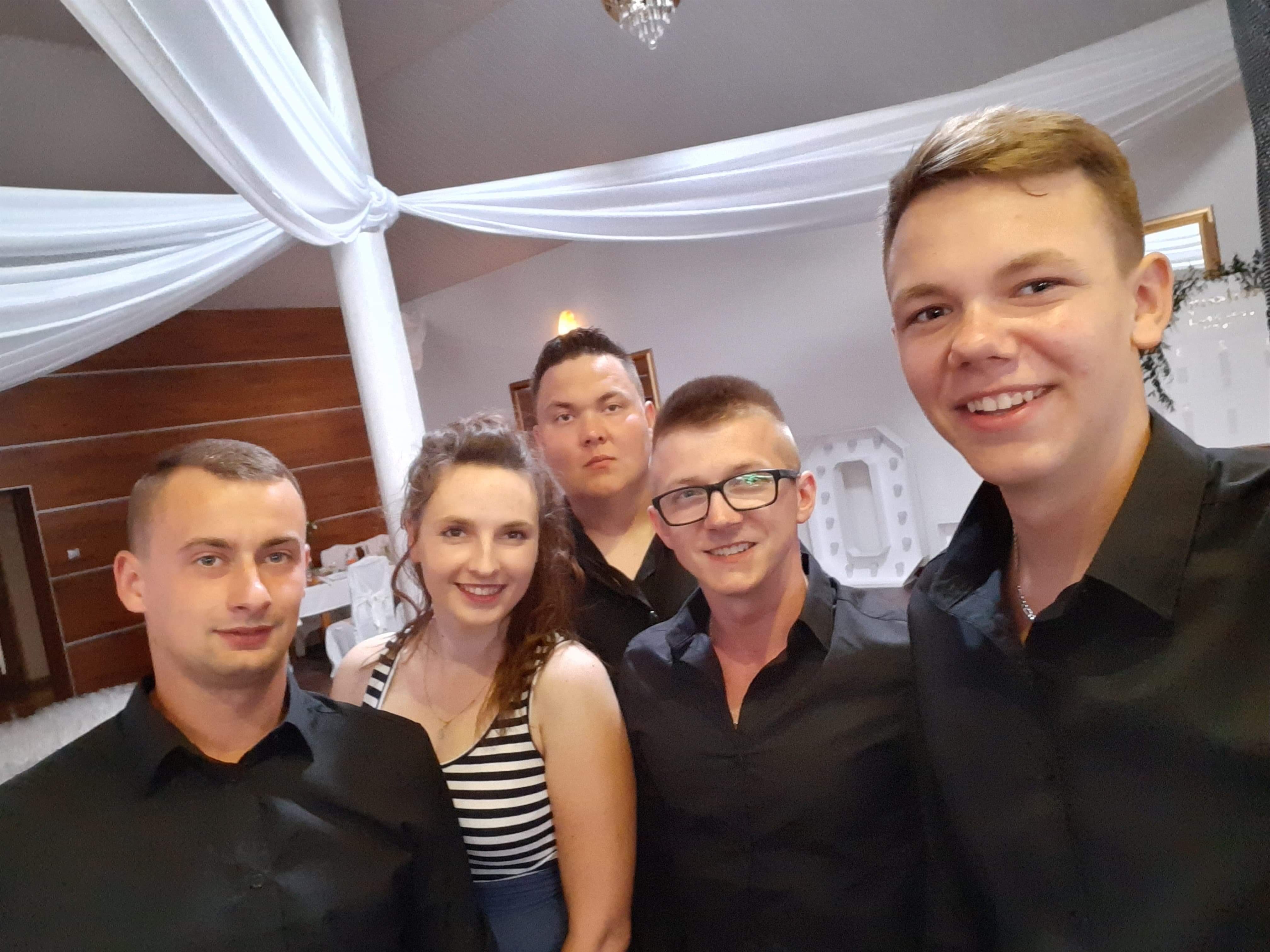 Pięcioosobowa grupa młodych ludzi pozuje do zdjęcia na tle sali udekorowanej białymi draperiami i kryształowym żyrandolem.