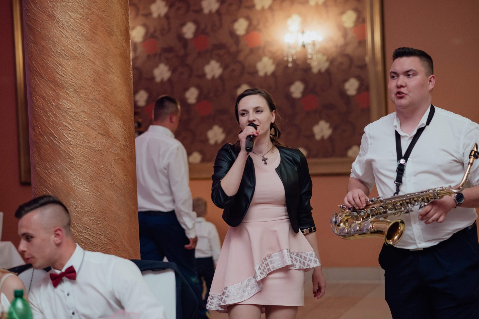 Wokalistka z mikrofonem i saksofonista w trakcie występu na tle kolumny w sali z wzorzystą tapetą.