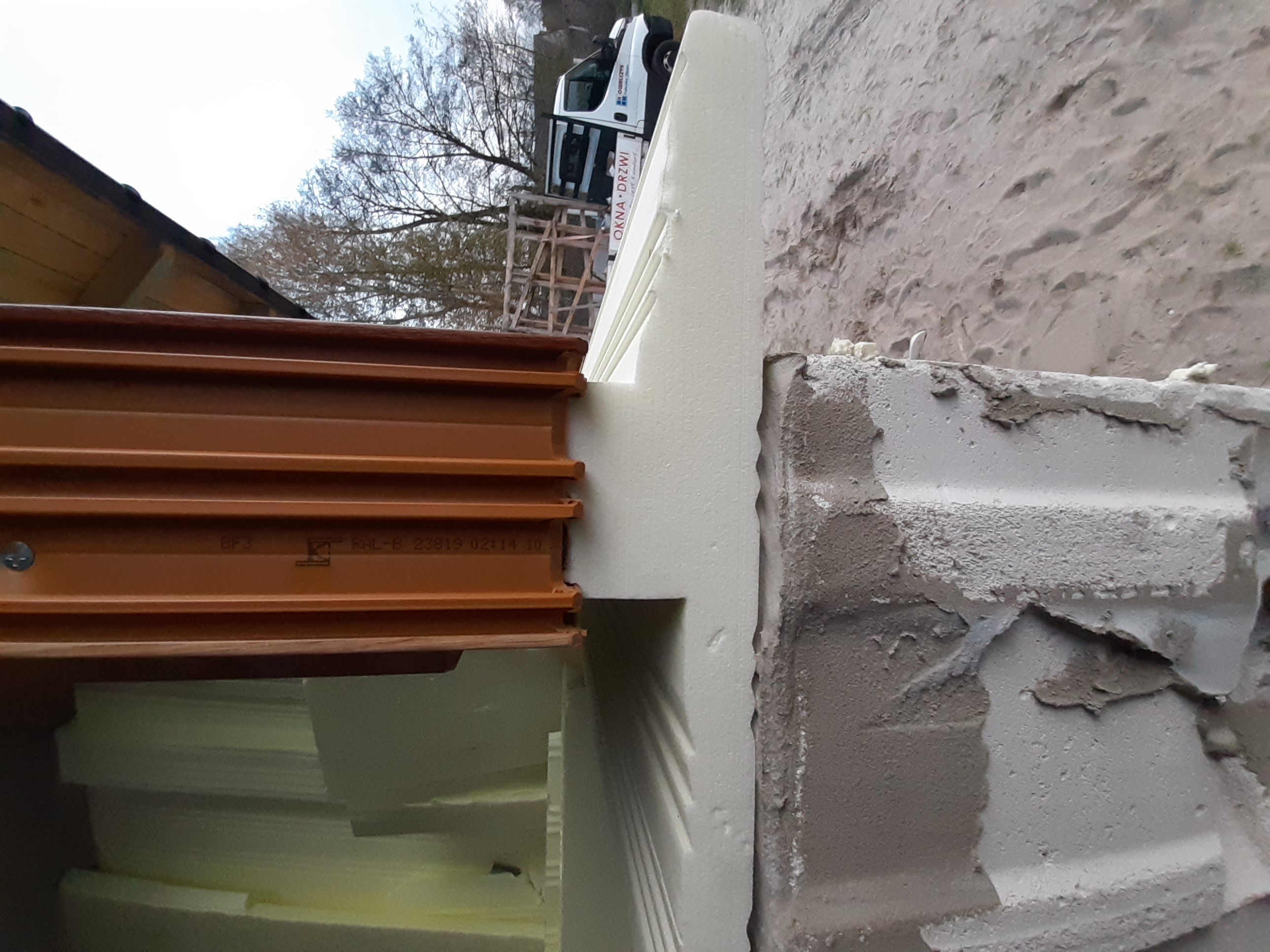 Montaż brązowego okna PCV z widocznym ociepleniem styropianowym i fragmentem ściany w surowym stanie, na zewnątrz widoczny samochód dostawczy z logo firmy.