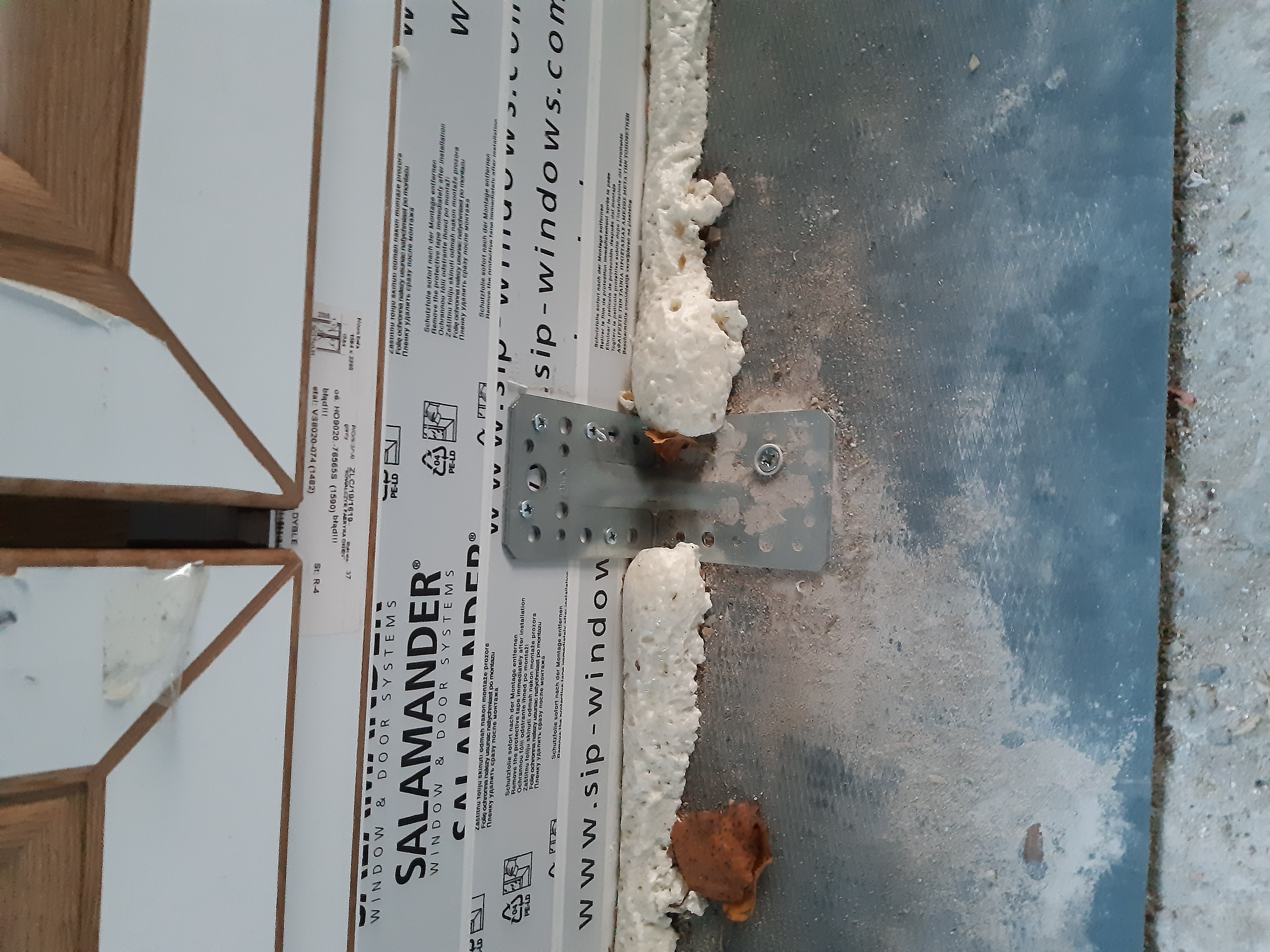 Detail montażu okna PCV z widoczną pianką montażową, metalowym mocowaniem i folią ochronną Salamander.