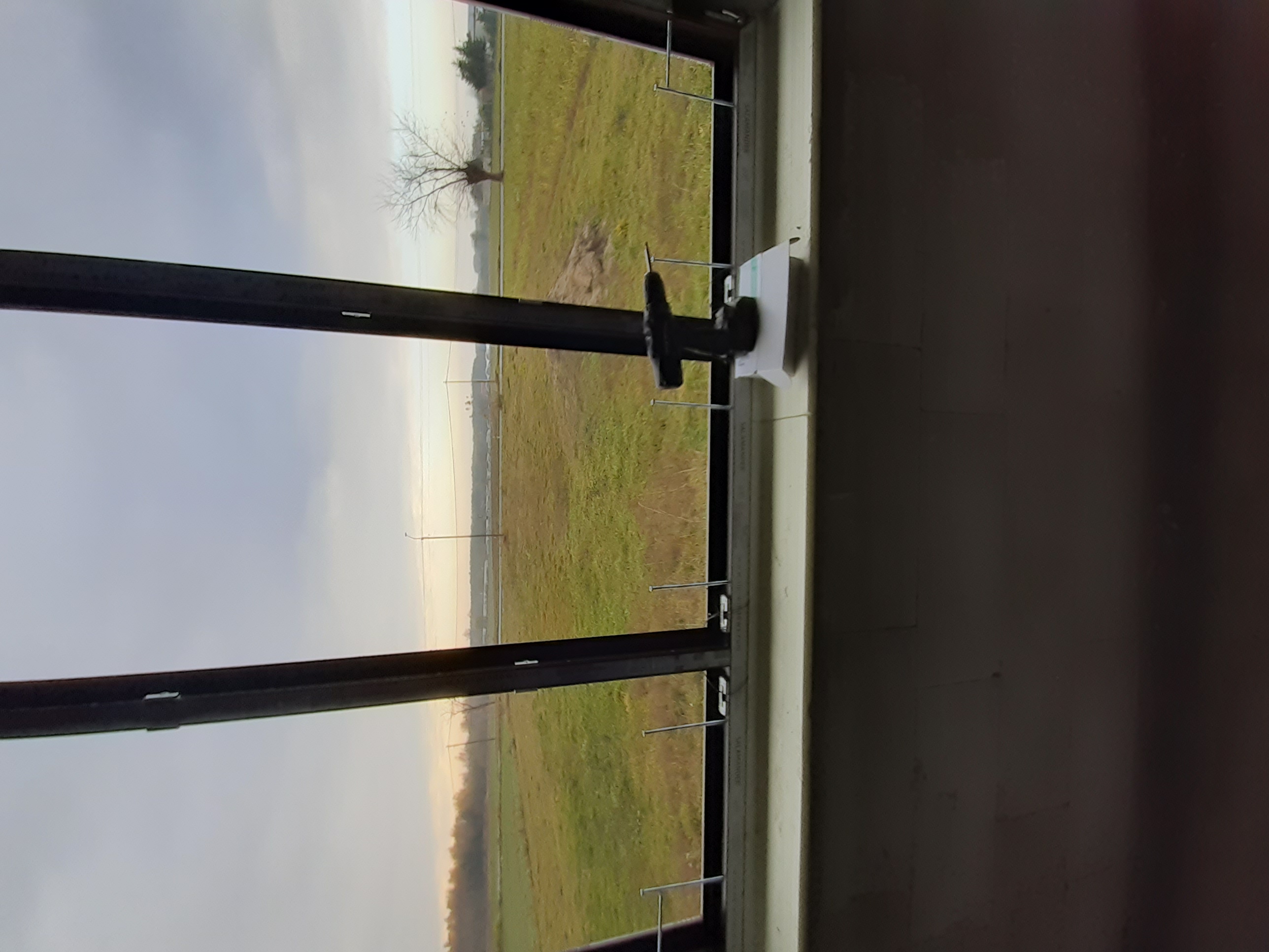 Widok przez okno PCV w trakcie montażu, widoczne mocowania i krajobraz za oknem z samotnym drzewem na łące.