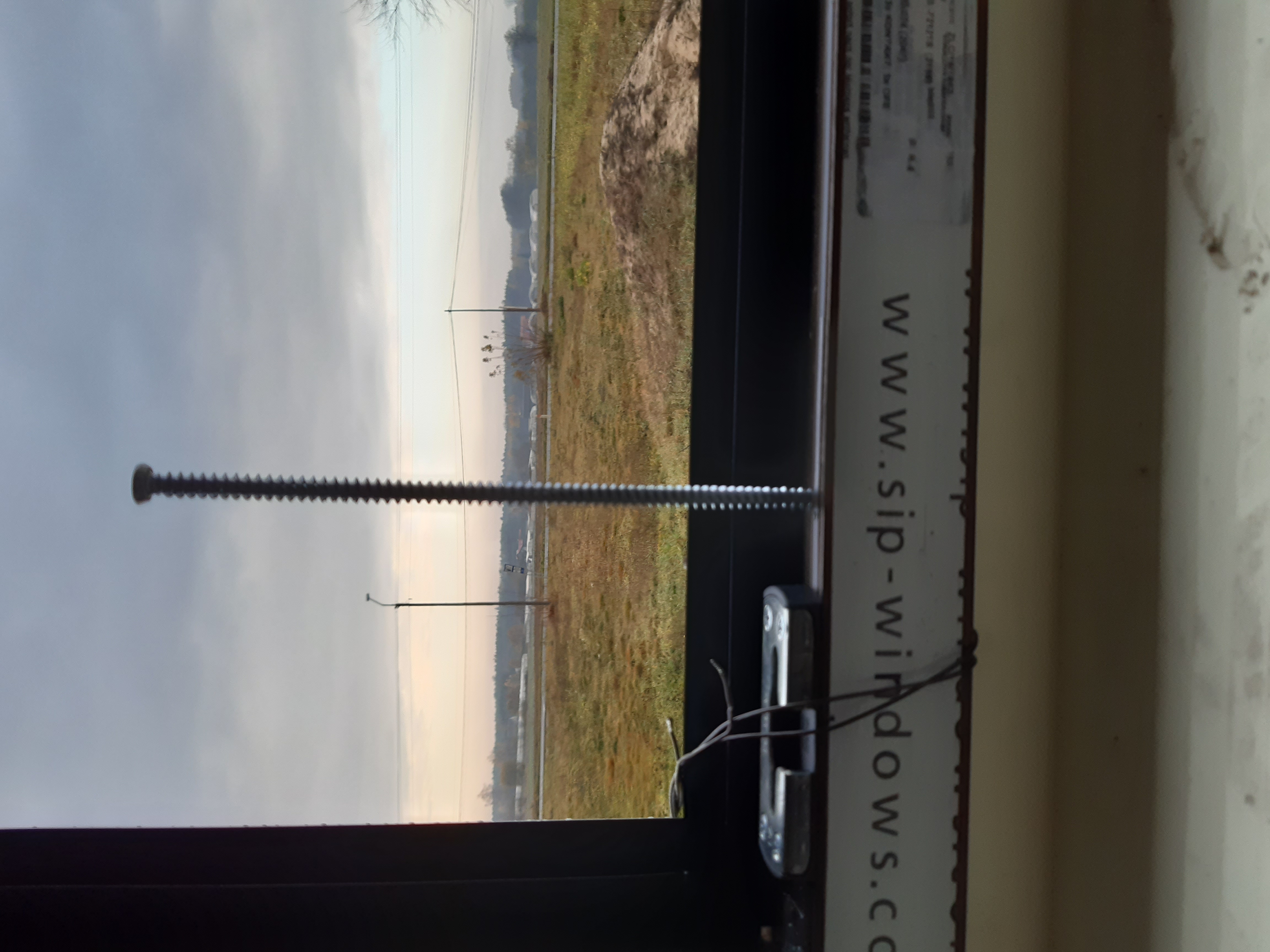 Widok z wnętrza okna na zewnątrz, widać wkręt w ramie okiennej oraz fragment krajobrazu za oknem.