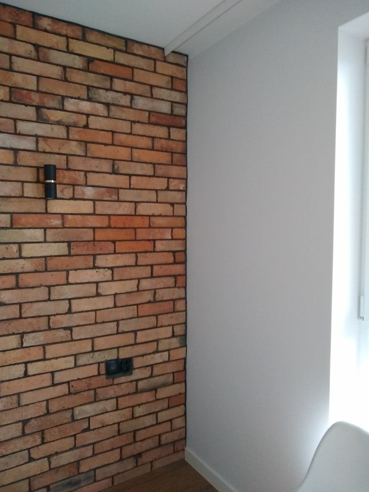 Ściana z cegły klinkierowej z czarnym kinkietem i gniazdkiem elektrycznym, kontrastująca z gładką, białą ścianą obok okna.