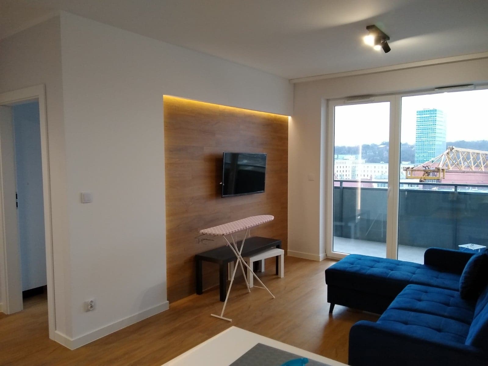 Nowoczesny salon z drewnianą ścianą podświetloną LED, telewizorem, deską do prasowania i niebieską sofą narożną z widokiem na miasto z balkonu.