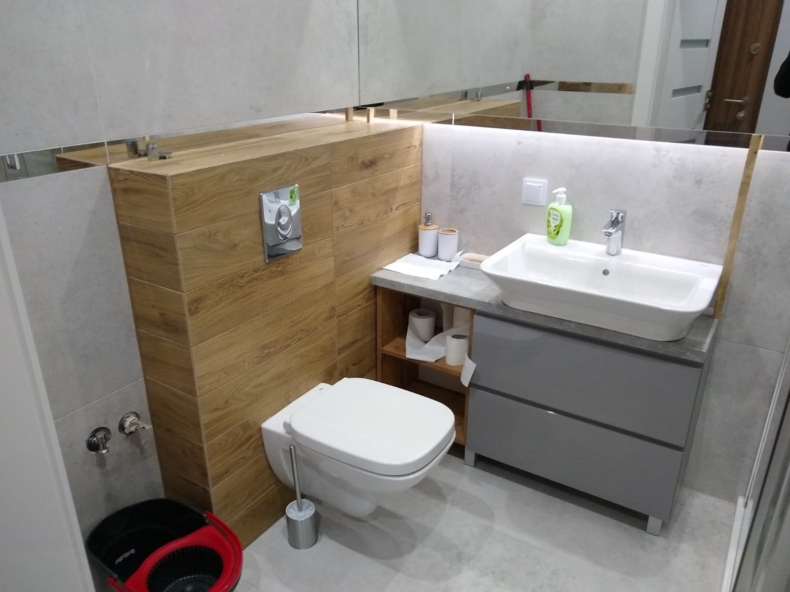 Nowoczesna łazienka z podwieszaną toaletą, zabudową z płytek imitujących drewno, szarą szafką pod umywalką i dużym lustrem na ścianie.