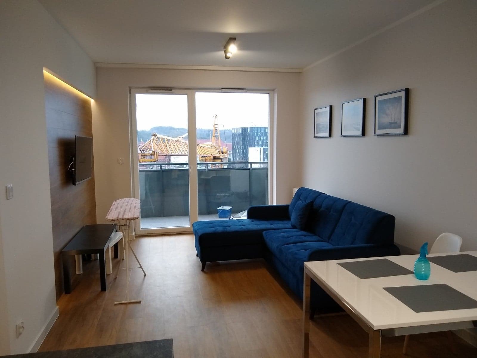 Salon po remoncie z niebieską sofą narożną, stołem z białym blatem i widokiem na budowę za oknem balkonowym.